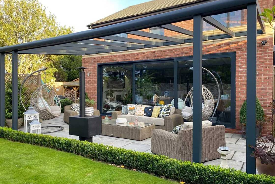 glass-roof-veranda-with-table-and-chairs Anthracite aluminium veranda and patio seating area with wood burner installation by Suffolk Outdoor Living in a landscaped garden.